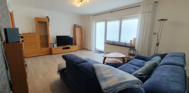 BookNoFees: Ferienwohnung in Langelsheim – Wohnzimmer mit blauem Sofa, Holzmöbeln und einer Balkontür